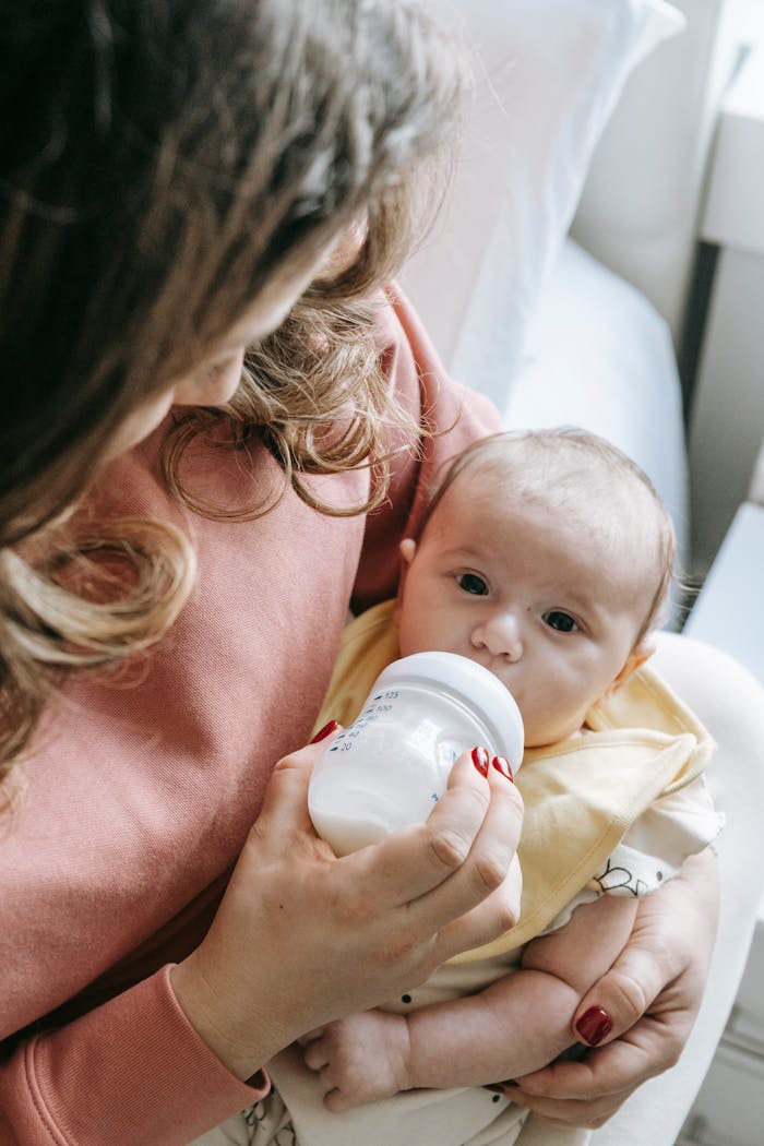 creative High angle of crop unrecognizable young mother in casual clothes sitting on bed and feeding adorable baby with milk from bottle in sunny morning